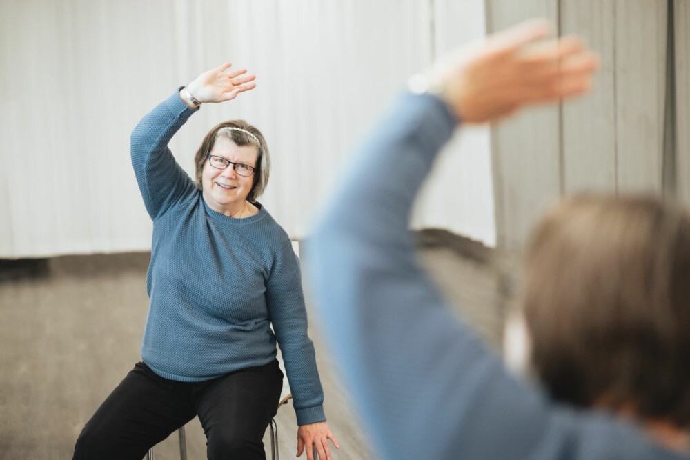 Kuvituskuva. Iäkäs nainen istuu tuolilla ja venyttää toista kättään korkealle.